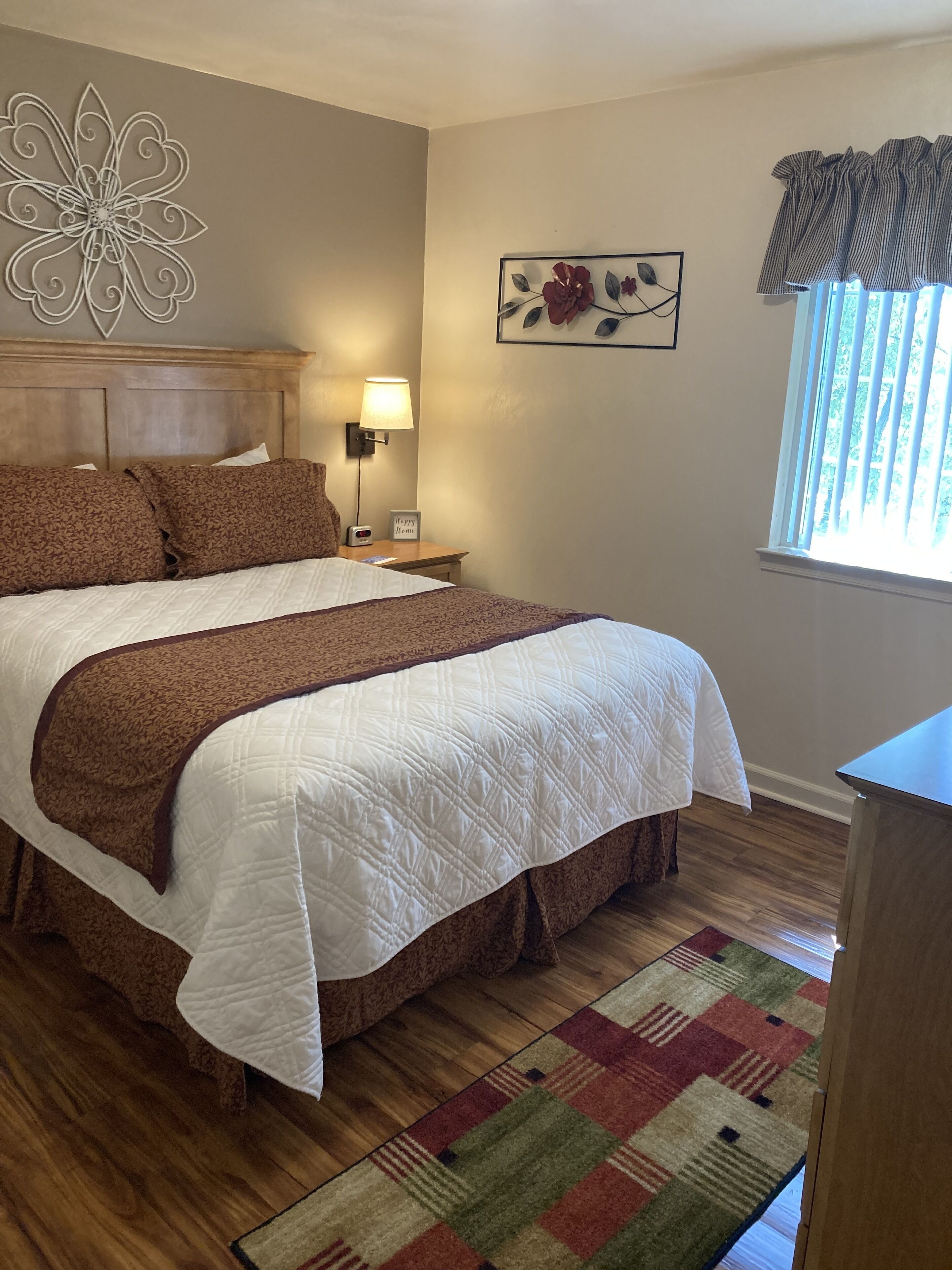 A neatly made bed with a brown and white quilt, a wooden headboard, a bedside table with a lamp, a framed picture on the wall, and a window with blinds.