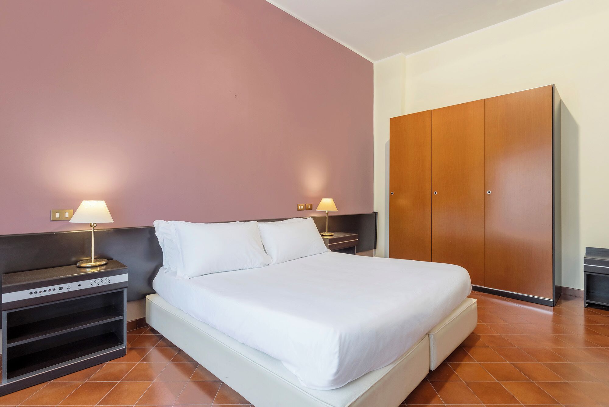 A hotel room with a bed, two bedside tables with lamps, a wardrobe, and a built-in TV stand.