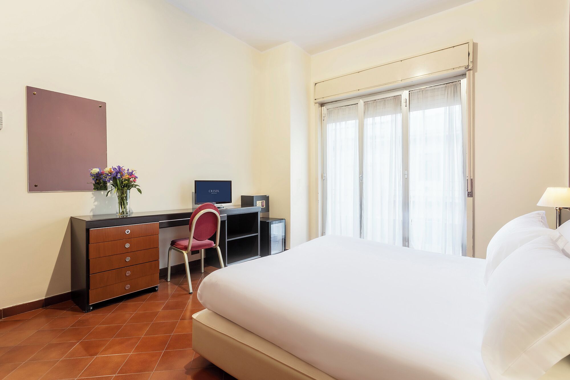 A hotel room with a bed, a desk with a computer, a chair, a wardrobe, and a window with curtains.