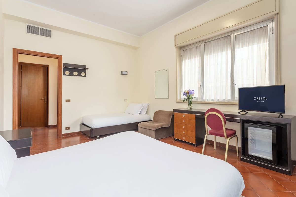 A hotel room with a bed, a desk with a computer, a chair, a small cabinet, and a window with curtains.