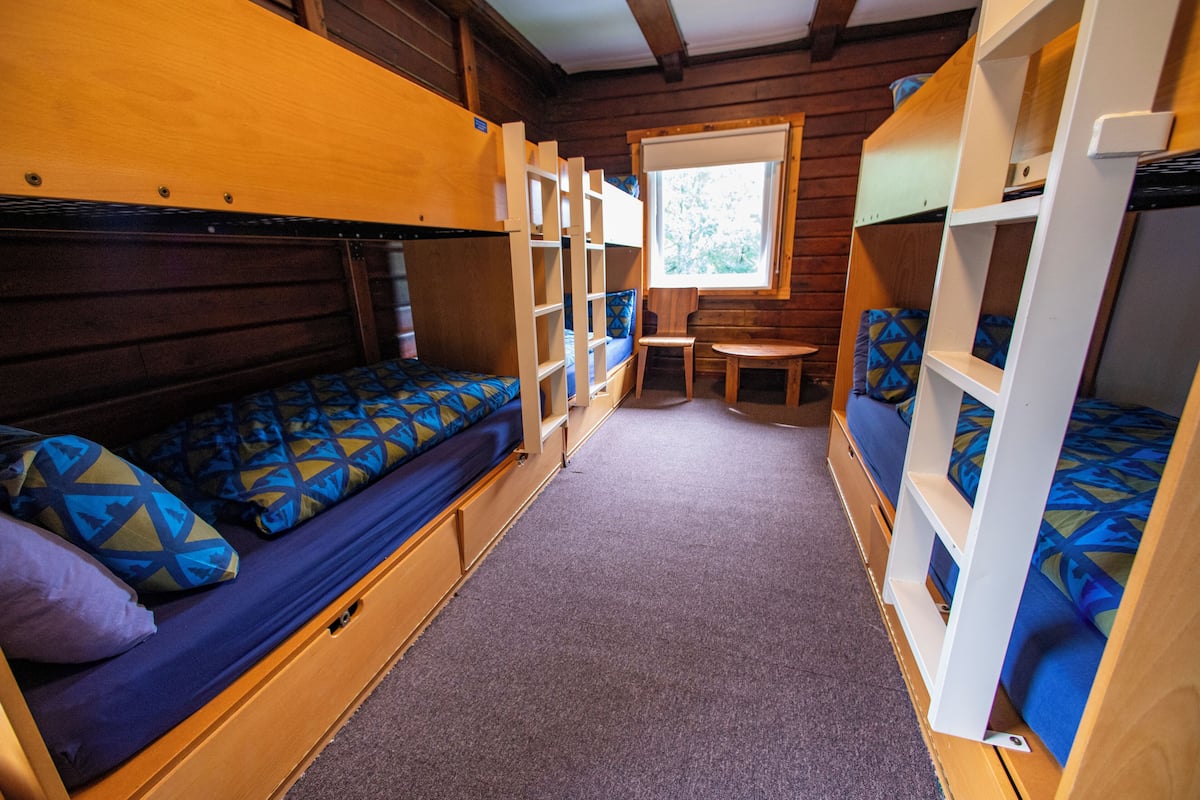 A compact, wooden cabin-style room with bunk beds, a small table, and a window providing natural light.