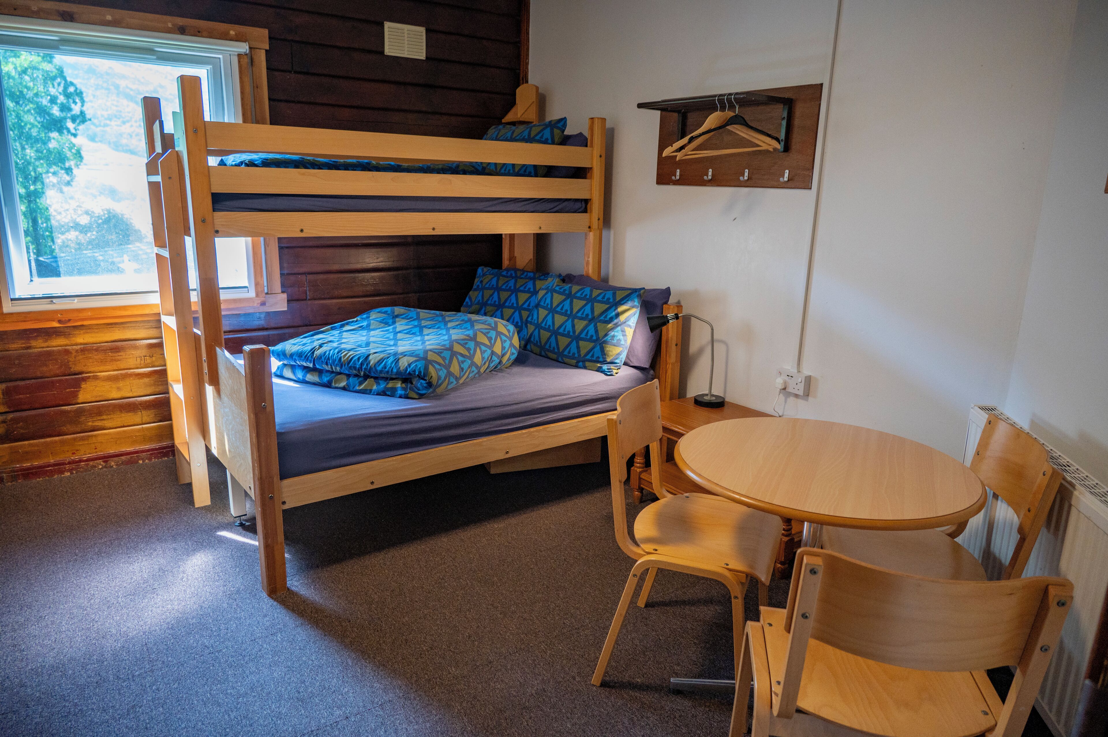 A bunk bed with a desk and chair, a window with a view of trees, and a wall-mounted coat rack.