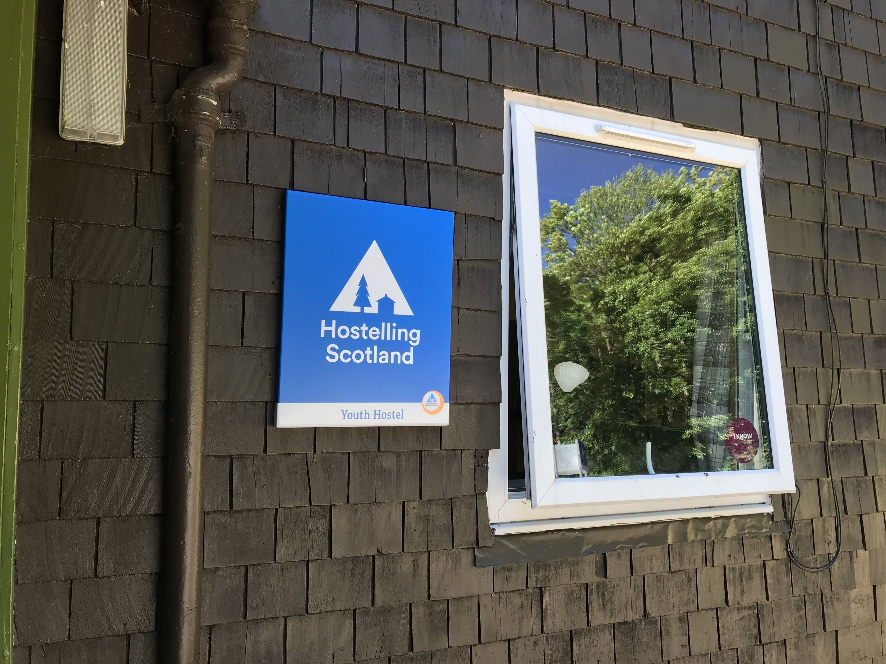 Une enseigne indiquant un auberge de jeunesse, avec un pictogramme représentant une montagne et une maison, une fenêtre donnant sur des arbres et un mur de briques.