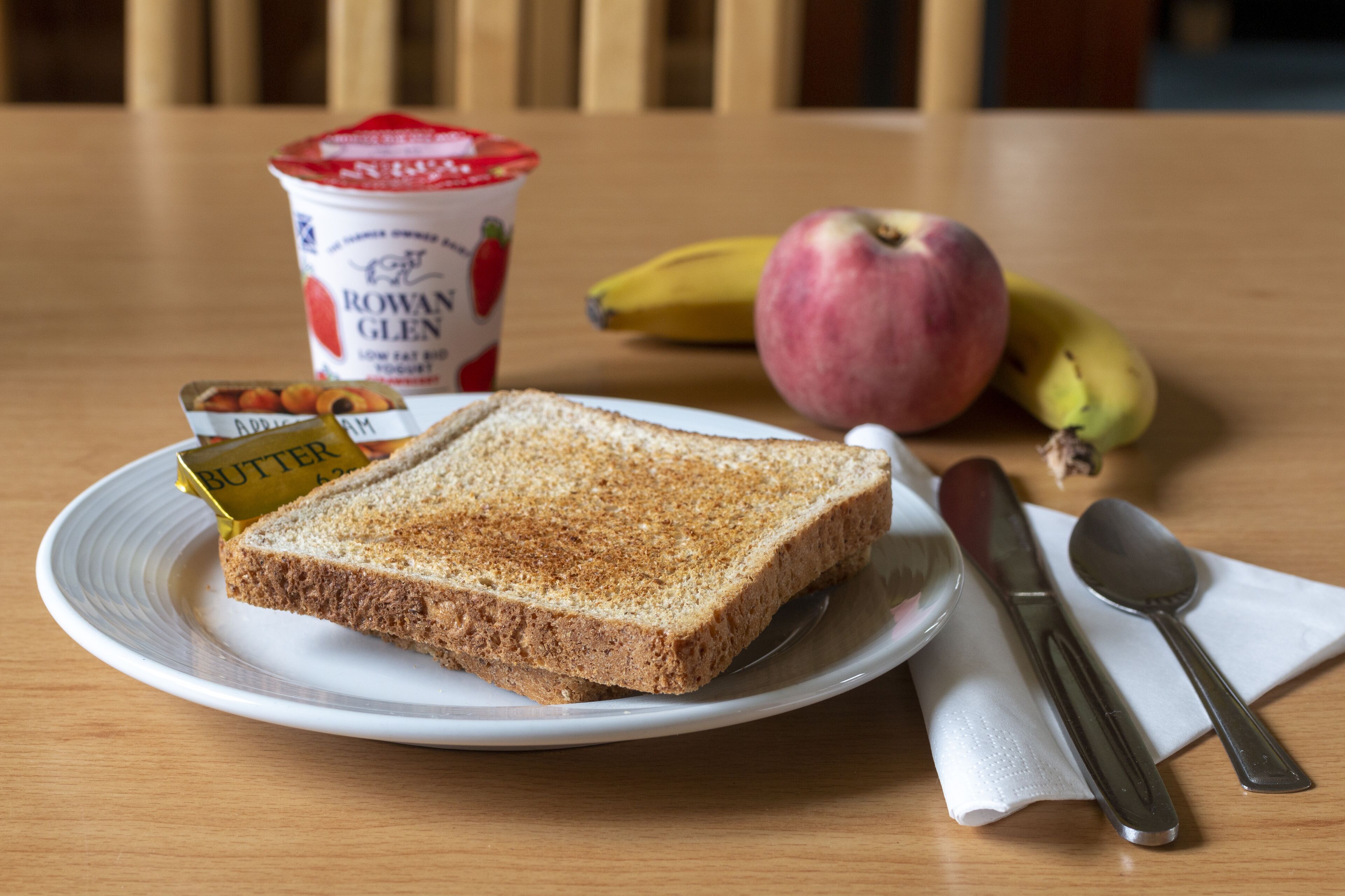 A plate with butter, a slice of bread, a banana, an apple, and a container of yogurt on a wooden table.