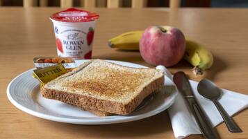 A plate with butter, a slice of bread, a banana, an apple, and a container of yogurt on a wooden table.