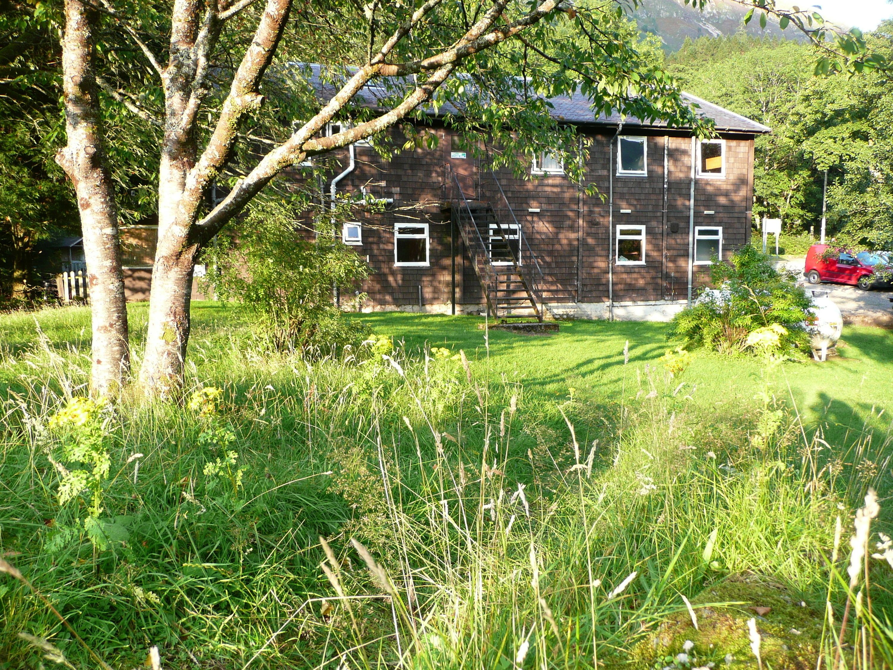 A rustic two-story building with a brick exterior, surrounded by lush greenery and trees.