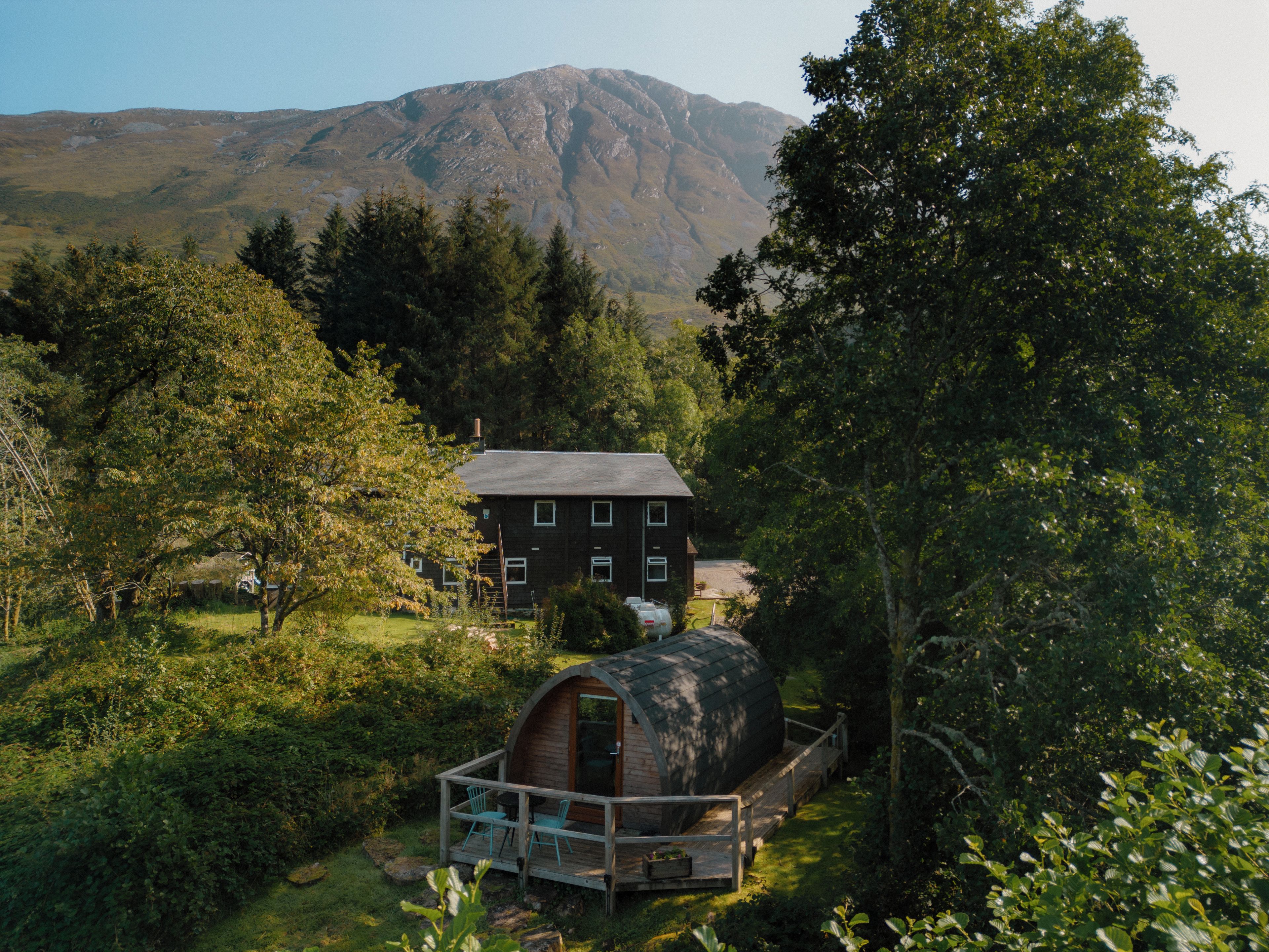 A cabin with a deck and a barrel-shaped structure in a forested area with a mountain in the background.