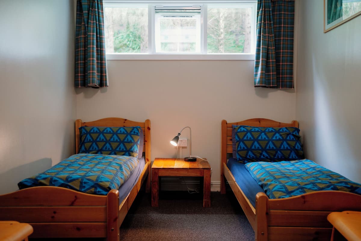 A room with two wooden beds, a small table with a lamp, and a window with curtains.