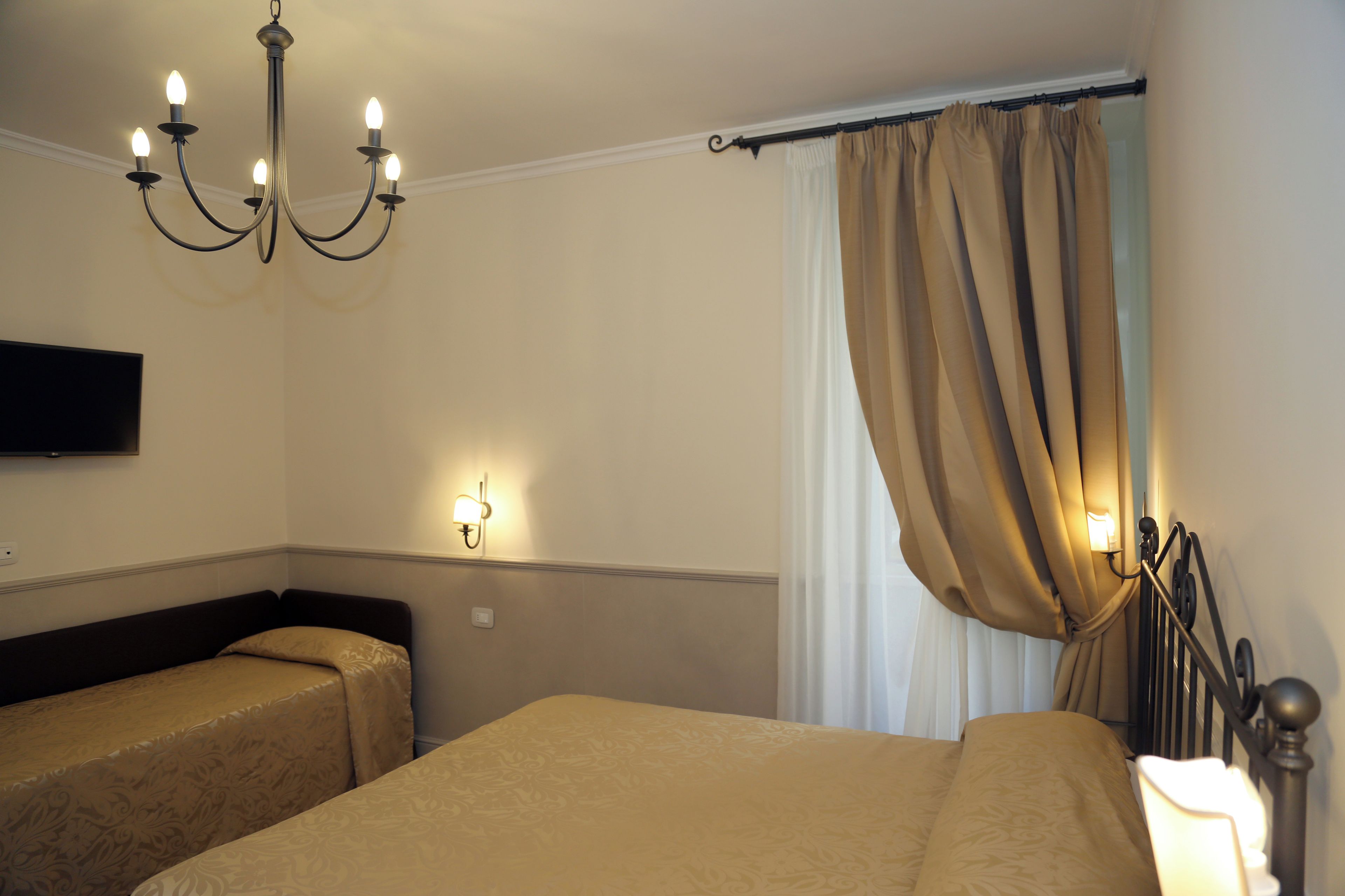 A hotel room with two single beds, a chandelier, a wall-mounted TV, and a window with curtains.