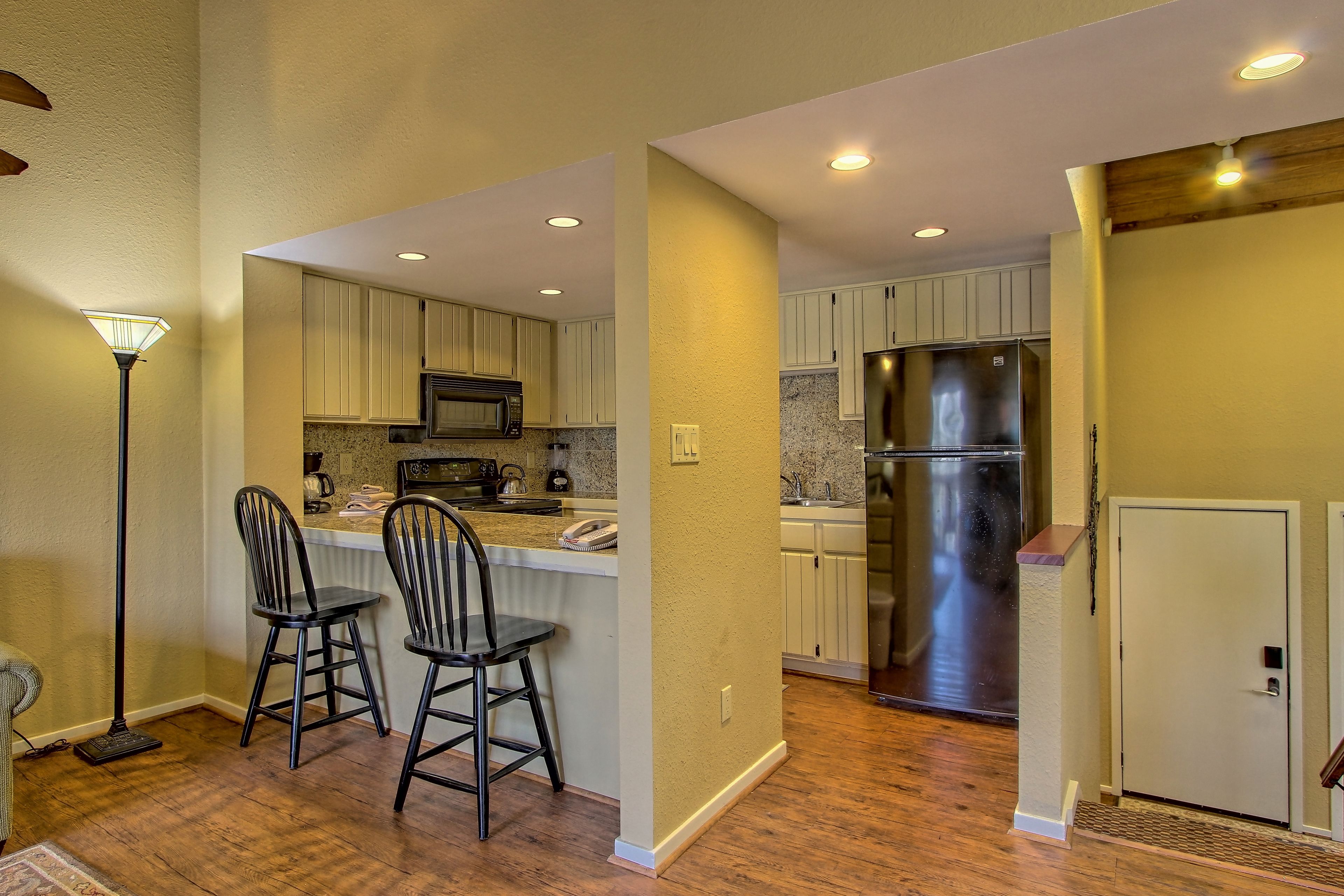 A modern kitchen with wooden flooring, white cabinets, a stainless steel refrigerator, and two bar stools.