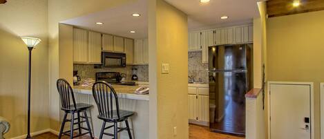 A modern kitchen with wooden flooring, white cabinets, a stainless steel refrigerator, and two bar stools.