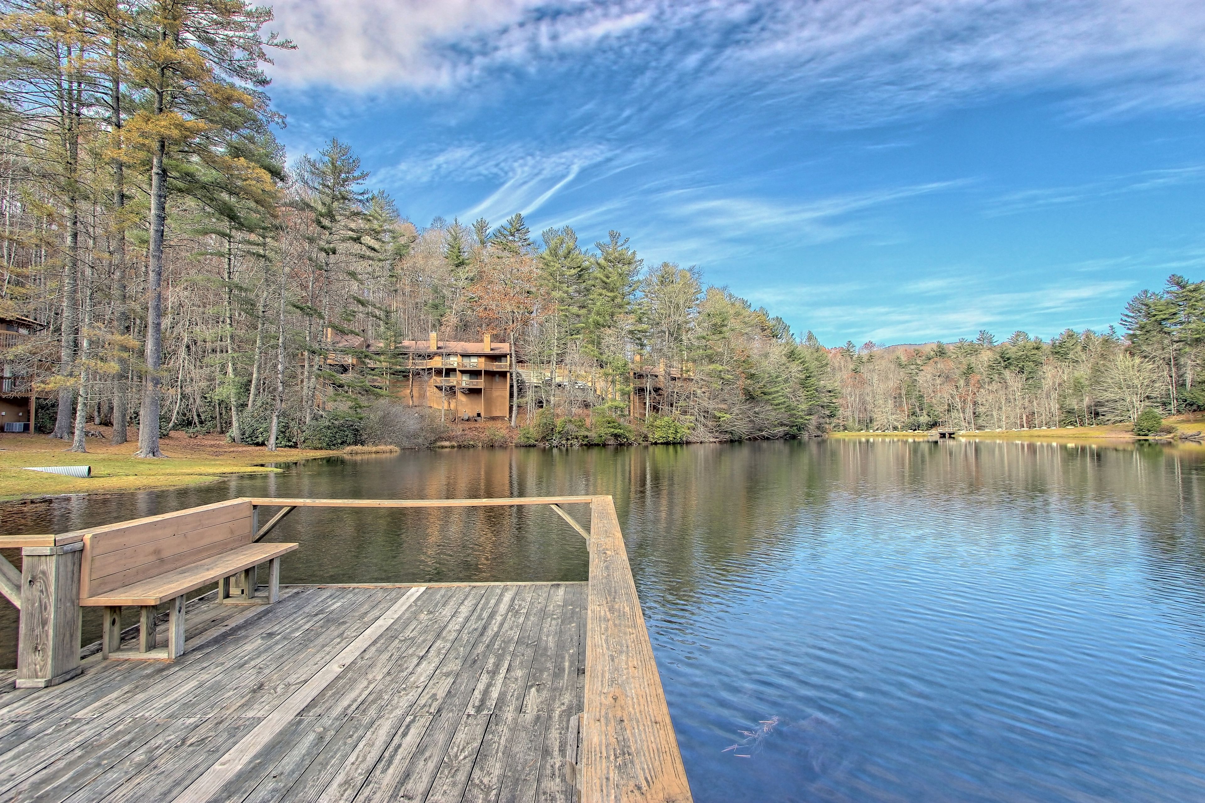 A wooden dock extending into a calm lake, surrounded by trees and a building in the background.