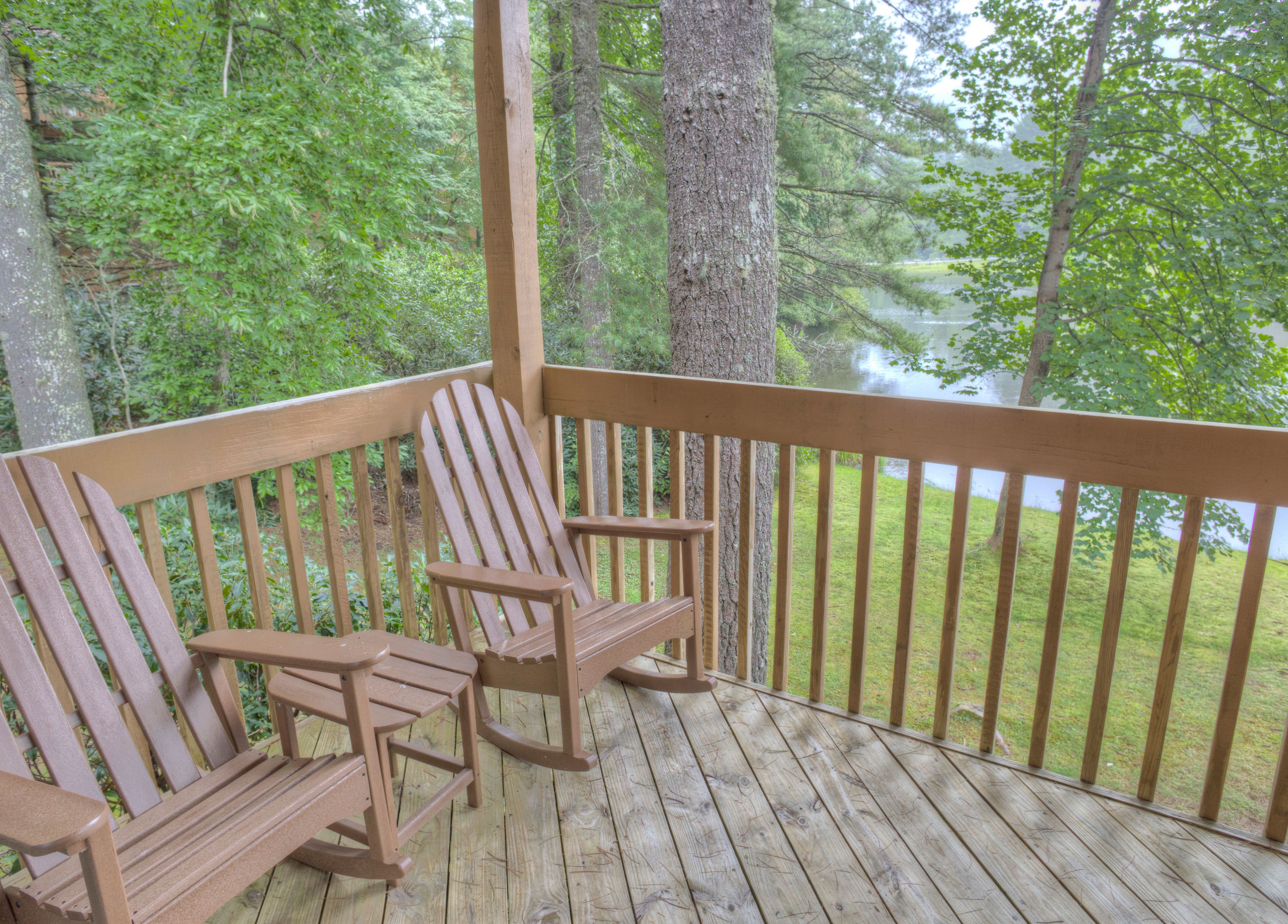 Una terraza de madera con dos sillas Adirondack mirando un lago, rodeado de árboles.