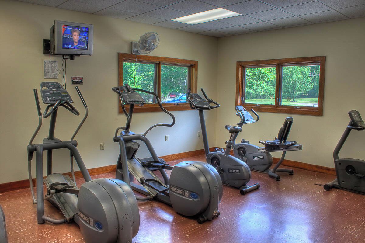 A gym with several stationary bikes and treadmills, large windows with a view of trees, and a television mounted on the wall.