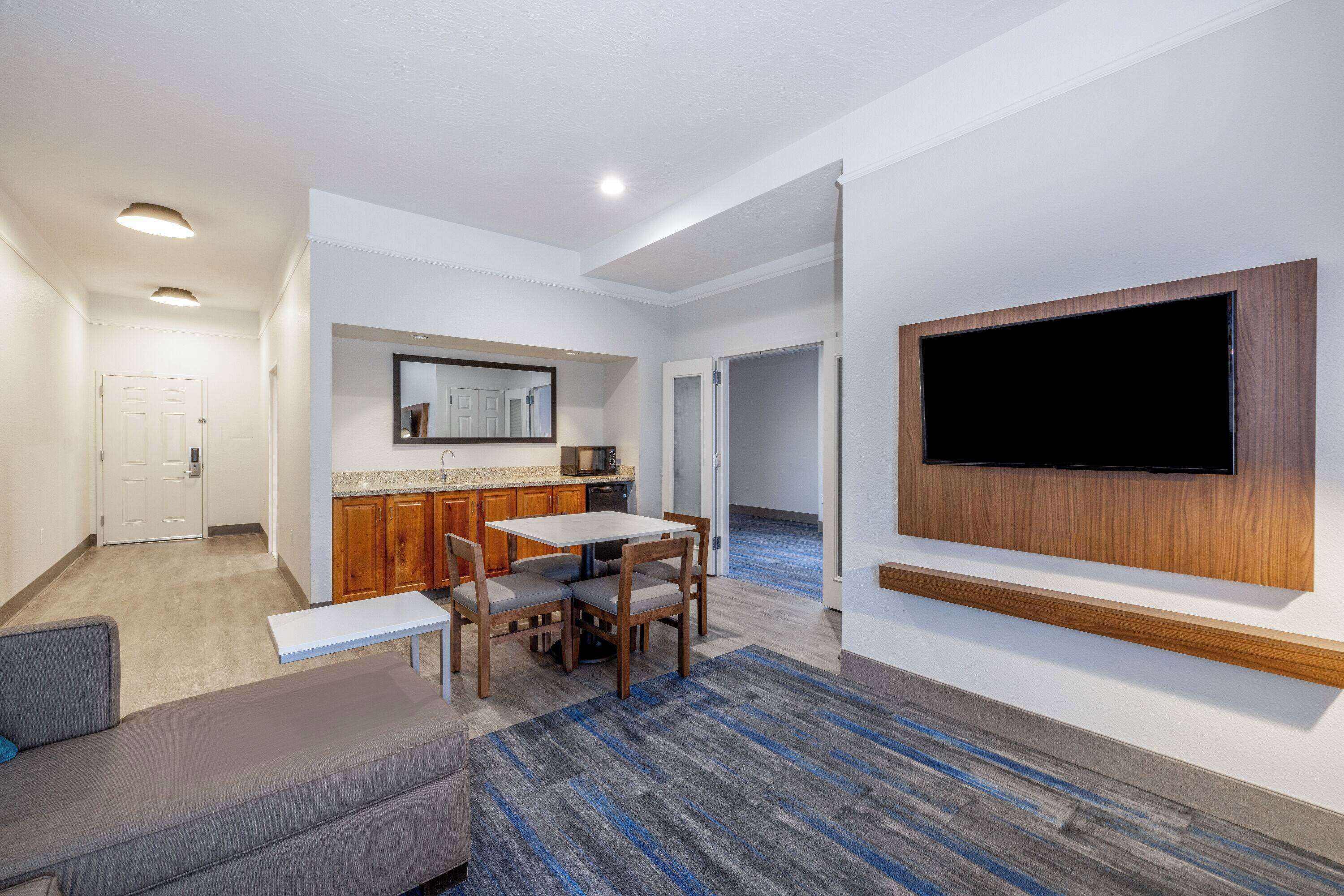 A modern living room with a flat-screen TV mounted on the wall, a dining area with a table and chairs, and a kitchen area with wooden cabinets and a microwave.