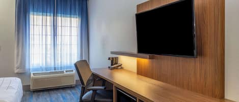 A hotel room with a bed, a desk with a chair, a flat-screen TV mounted on the wall, and a window with curtains.