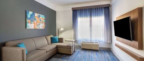 A modern hotel room with a sectional sofa, a flat-screen TV, a large window with curtains, and a painting on the wall.