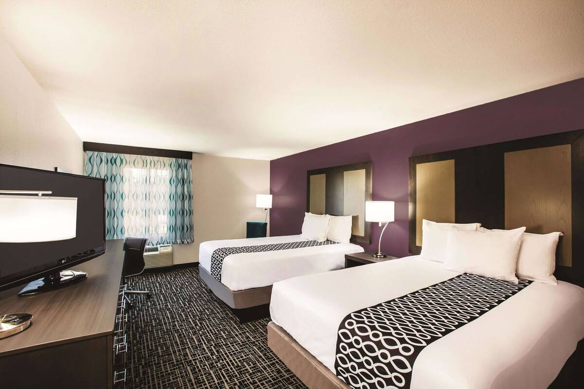 Habitación de hotel con dos camas, un escritorio con computadora, televisor y alfombra estampada.