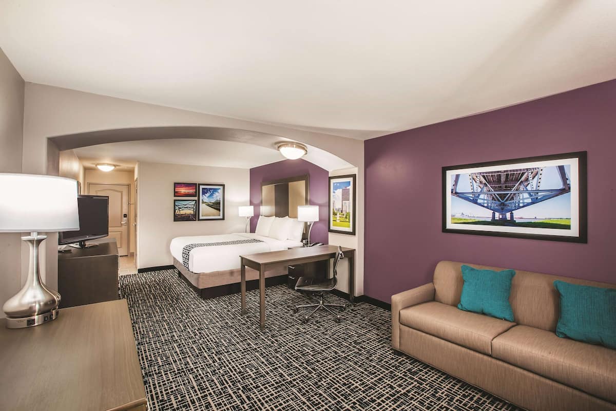 Habitación de hotel con cama, escritorio y sofá. Hay un televisor sobre un pedestal, una lámpara y un cuadro en la pared.