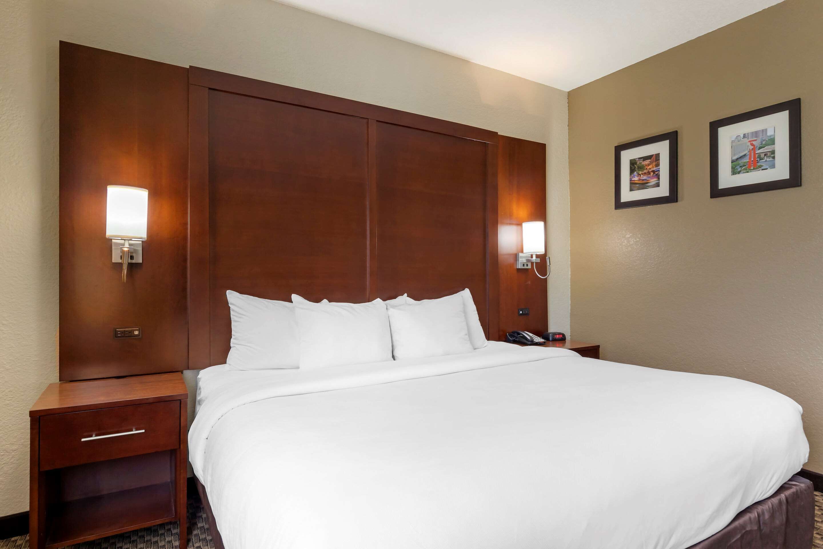 Una habitación de hotel con una cama grande, cabecera de madera, dos mesitas de noche con lámparas y dos cuadros en la pared.
