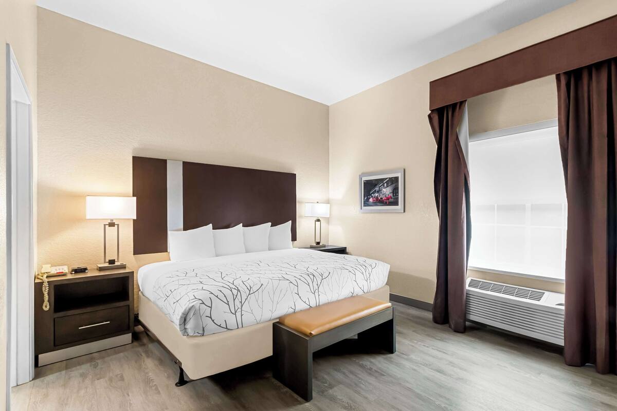 Une chambre d’hôtel avec un grand lit, des tables de chevet avec des lampes, un banc, une fenêtre avec des rideaux et un climatiseur fixé au mur.