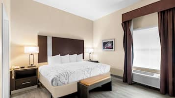 A hotel room with a large bed, bedside tables with lamps, a bench, a window with curtains, and a wall-mounted air conditioning unit.