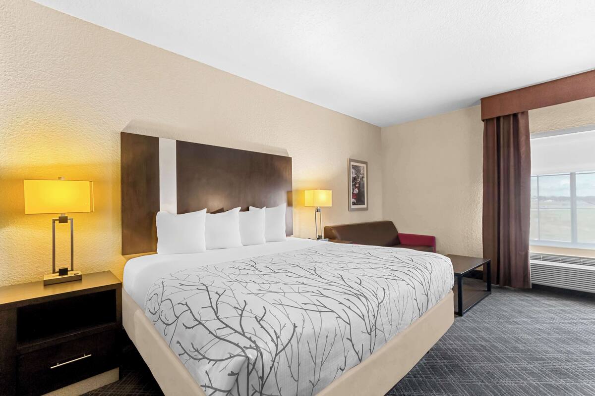 Une chambre d’hôtel avec un grand lit, deux tables de chevet avec des lampes, un fauteuil rouge, une petite table et une grande fenêtre avec des rideaux.