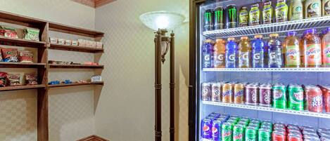 Rincón de una habitación de hotel con un refrigerador de bebidas repleto de diversas opciones, una lámpara de pie y estantes con aperitivos y artículos de tocador.