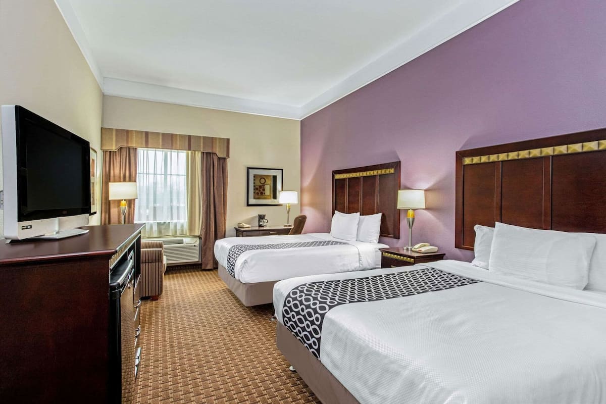 A hotel room with two beds, a TV, a desk, and a window with curtains.