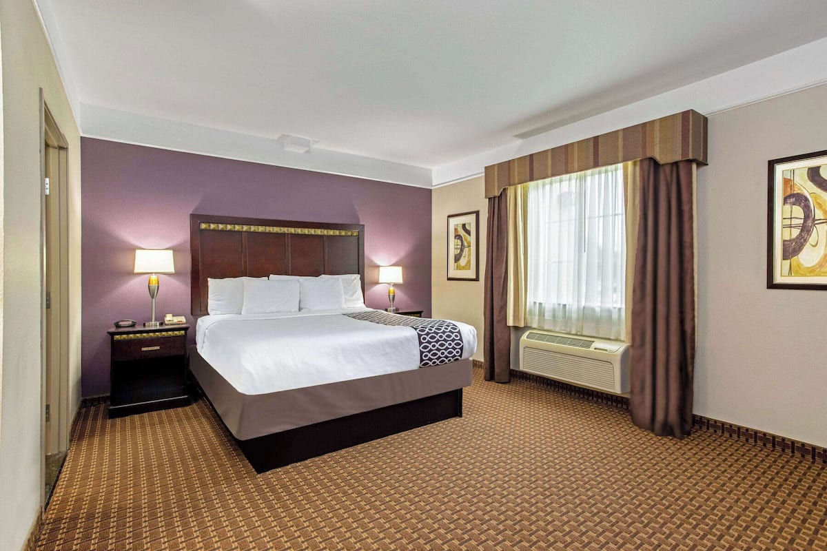 A hotel room with a large bed, two bedside tables with lamps, a window with curtains, and a wall-mounted air conditioner.