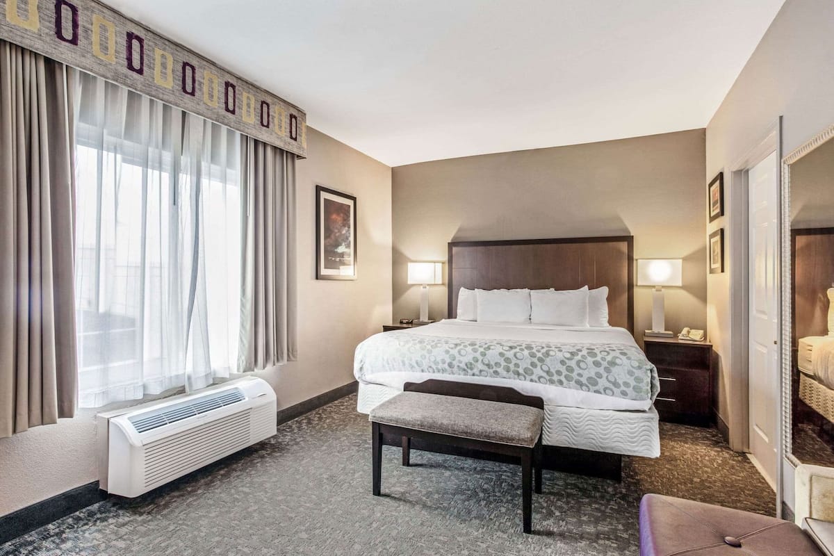 Une chambre d’hôtel avec un grand lit, des tables de chevet, un banc, un radiateur et une fenêtre avec des rideaux.