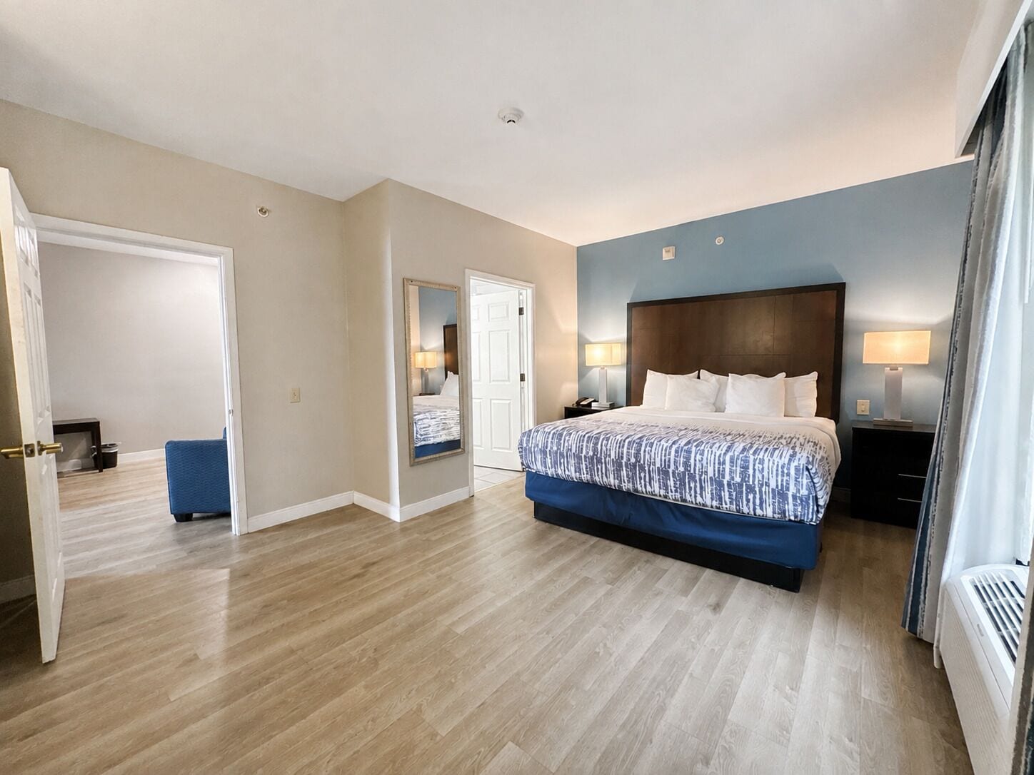 A hotel room with a large bed, bedside lamps, a door leading to another room, and a visible air conditioning unit.