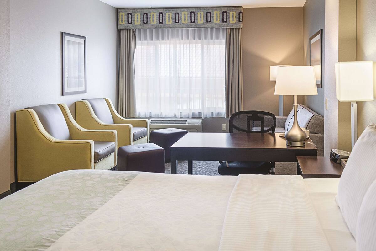 Une chambre d’hôtel avec un grand lit, deux fauteuils, une petite table, un bureau, une chaise, une lampe et une fenêtre avec des rideaux.