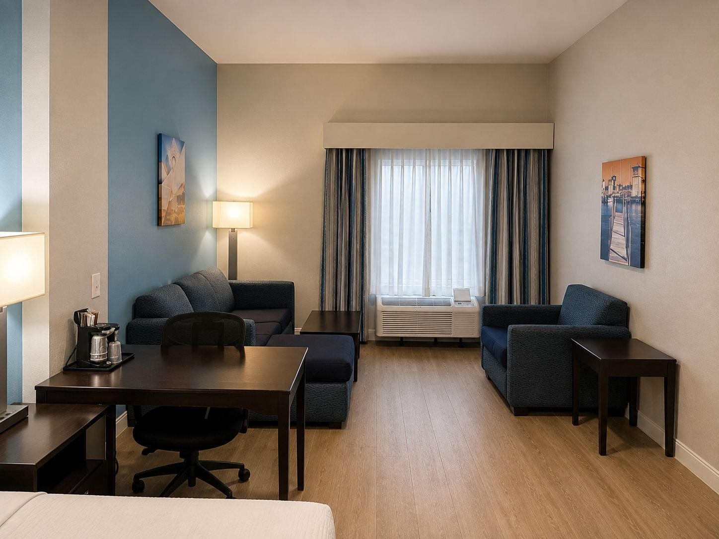 A hotel room with a desk, two couches, a chair, a lamp, and a window with curtains.