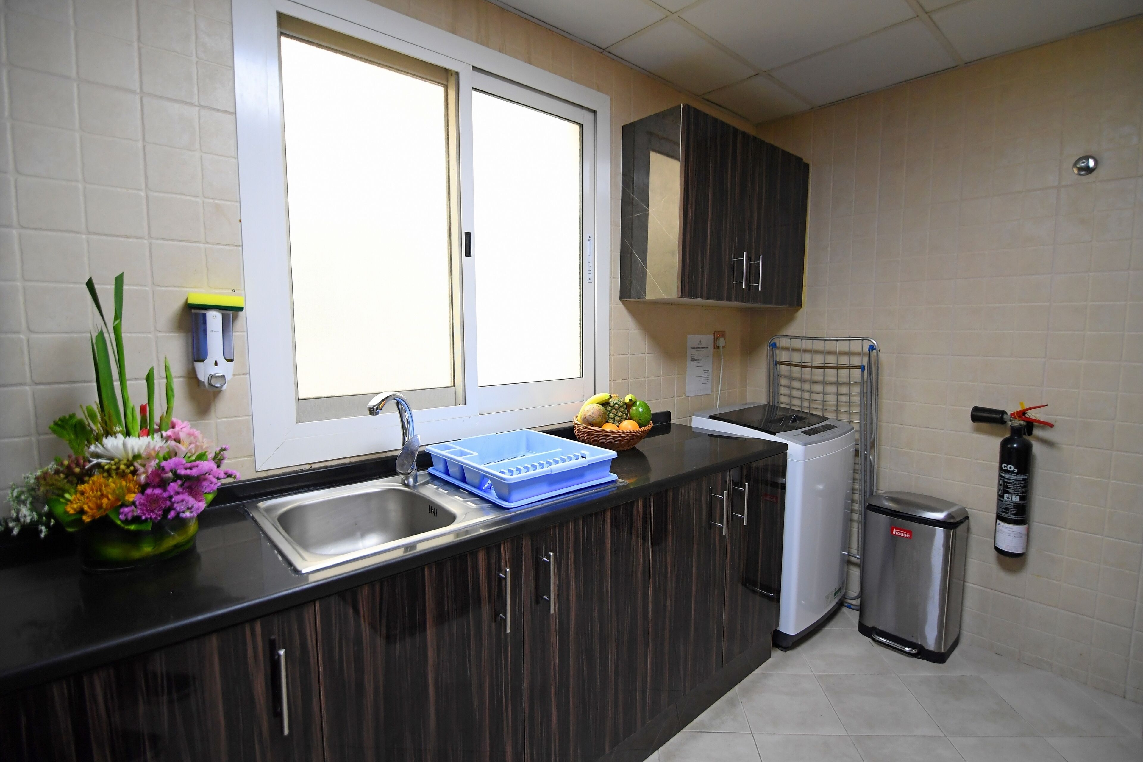 A kitchen with dark wooden cabinets, a stainless steel sink, a toaster, a trash bin, and a window with blinds.
