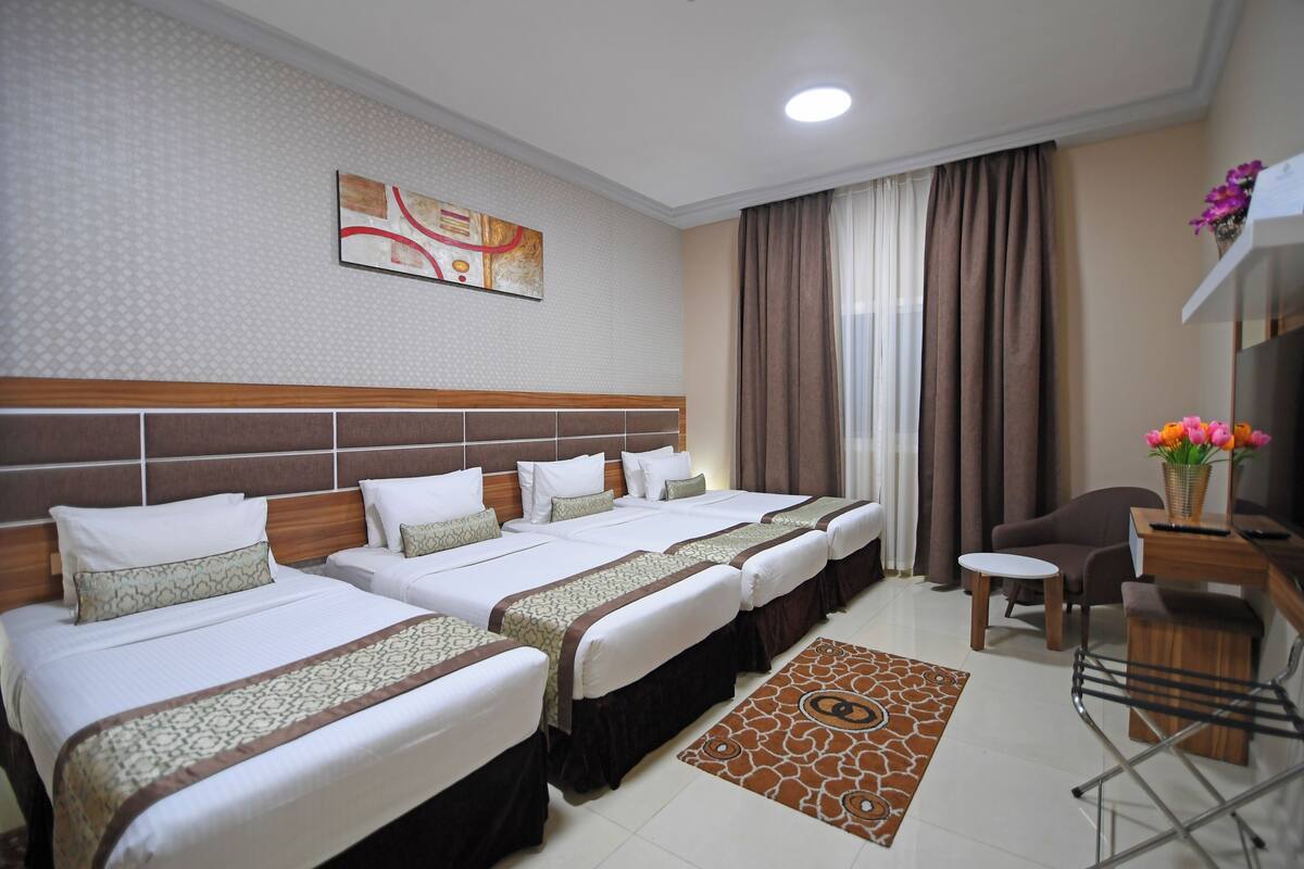 A hotel room with three beds, a desk, a chair, and a decorative rug.