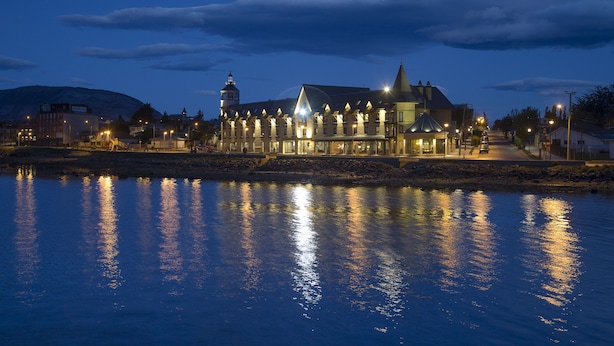 A waterfront hotel with a prominent tower and multiple lit windows reflecting on the water. - Hotel Costaustralis (Natales)