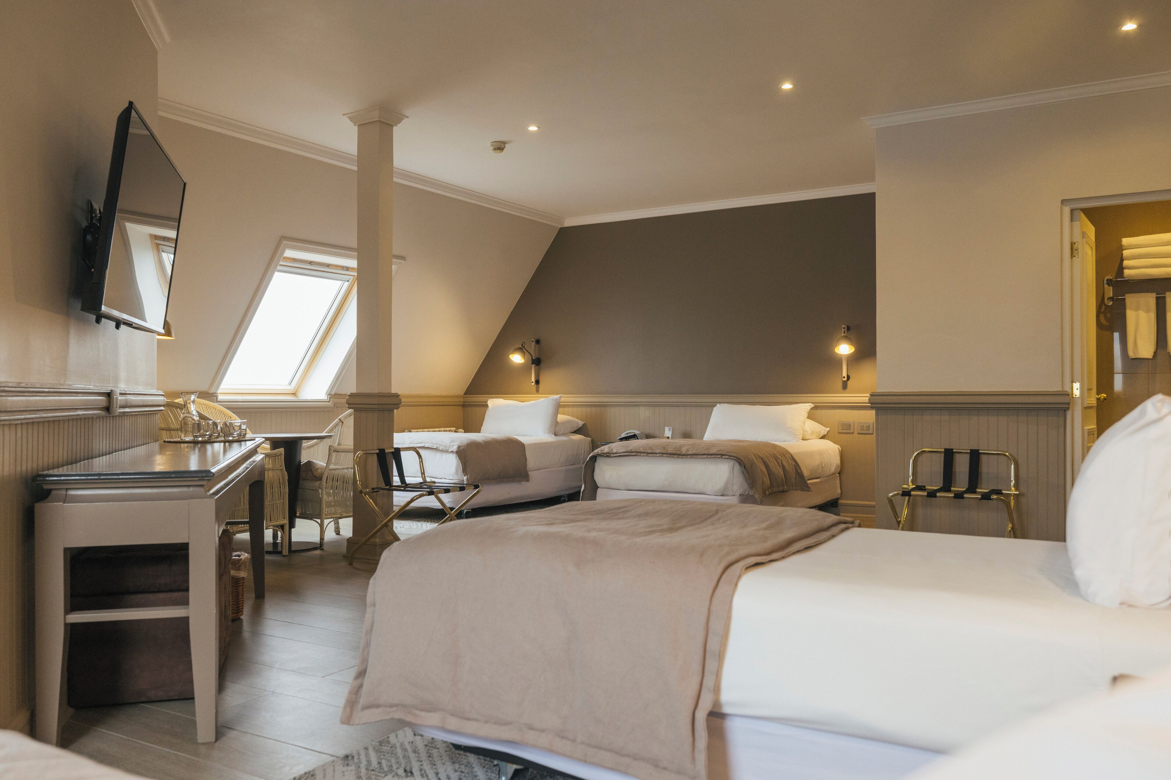 A hotel room with a large bed, a desk, and a chair. There is a window providing natural light and a mirror on the wall.