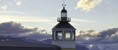 A lighthouse with a weather vane on top, overlooking a mountainous landscape and a body of water.