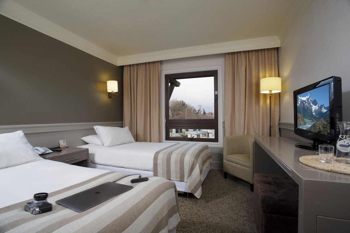 A hotel room with two beds, a desk with a television, and a view of the outdoors through a window.