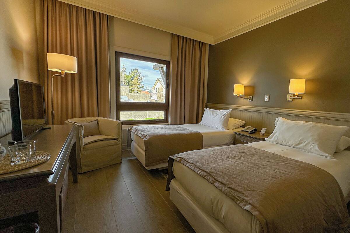 A hotel room with two beds, a desk with a chair, a TV, and a window with curtains.