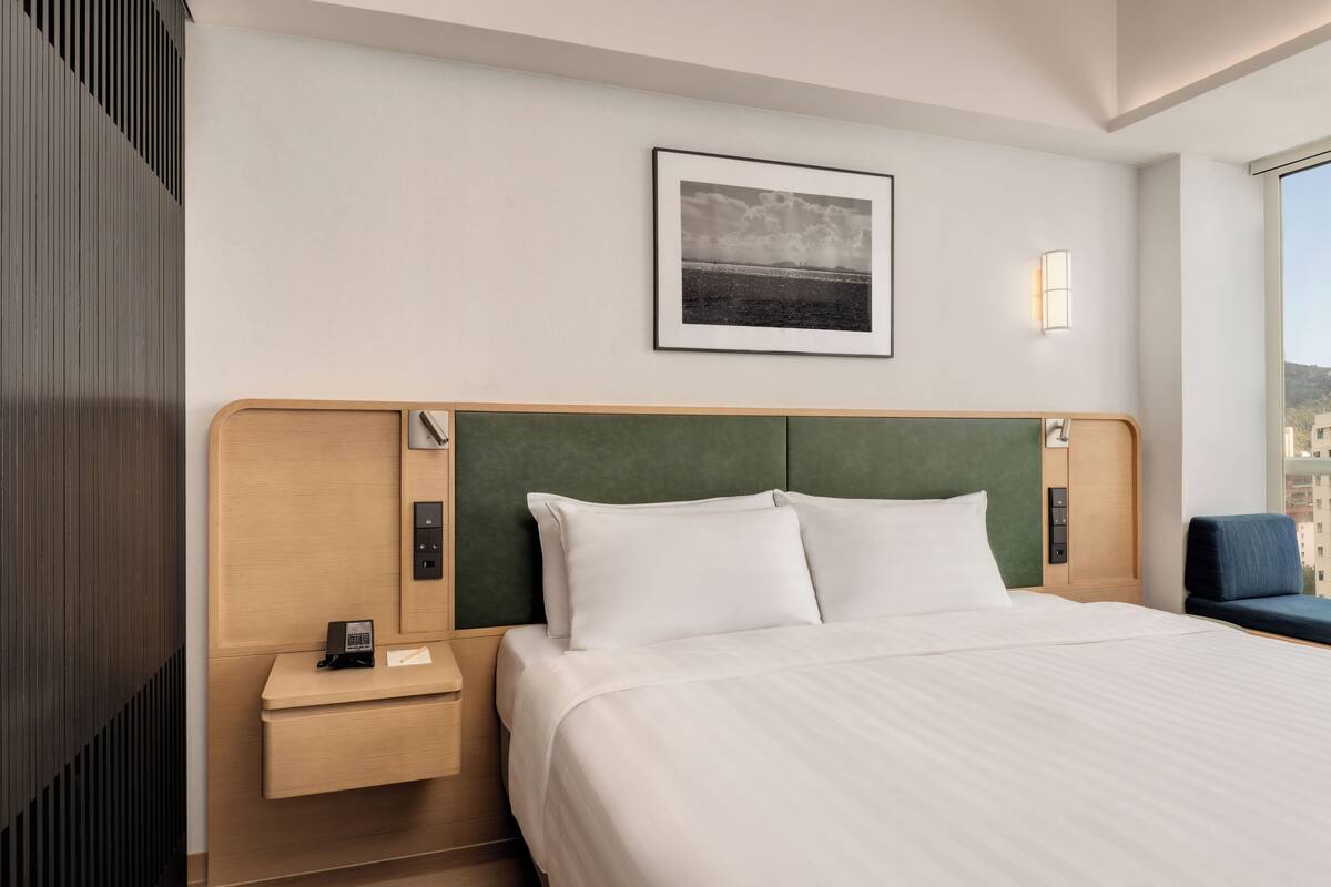 A hotel room with a bed, a nightstand, a phone, a framed picture on the wall, and a window with a view.