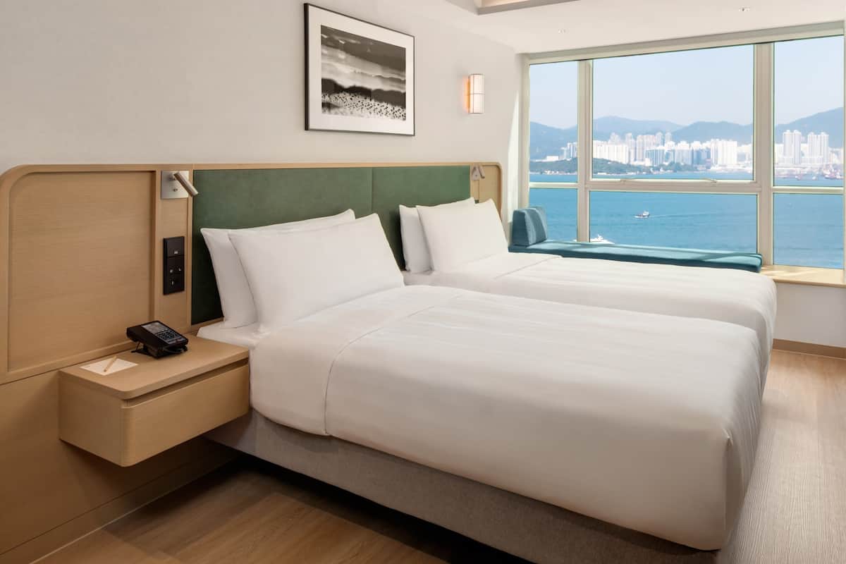 A hotel room with two beds, a nightstand with a phone, a window with a city view, and a framed picture on the wall.