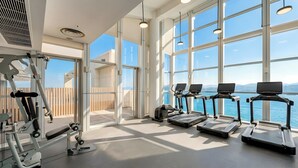 Ein Fitnessstudio mit Laufbändern, Kraftgeräten und großen Fenstern mit Blick auf ein Gewässer.