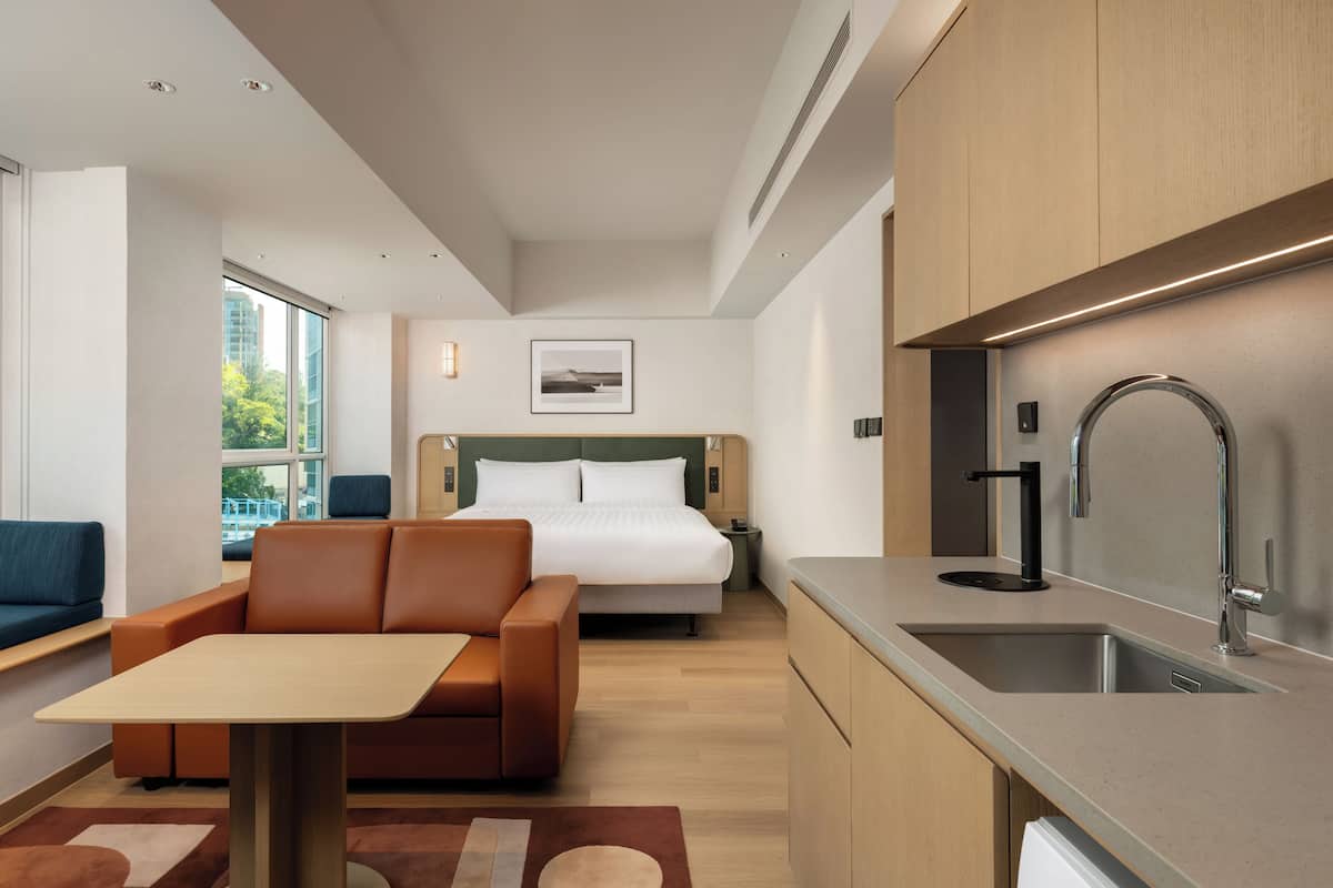 A modern hotel room with a bed, sofa, kitchenette, and a large window.
