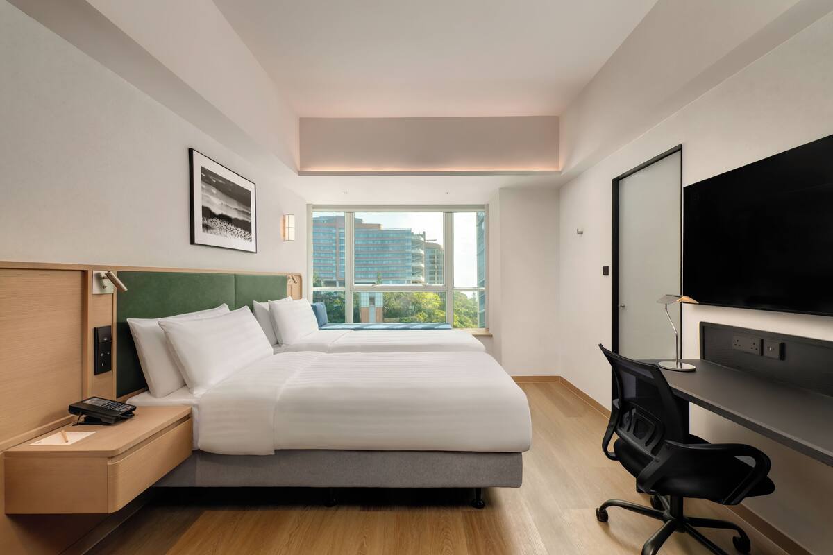 A hotel room with two beds, a desk, a chair, a TV, and a large window with a city view.