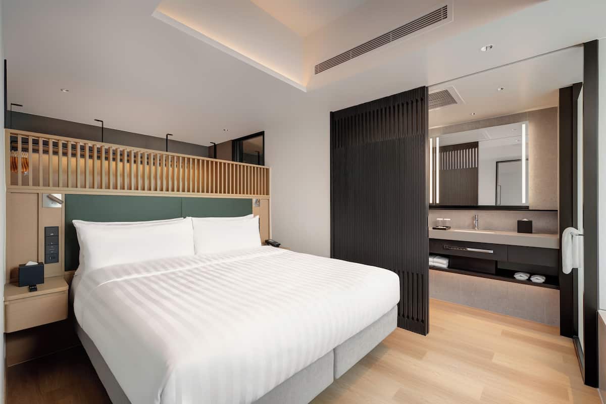 A modern hotel room with a large bed, a bedside table with a lamp, a built-in wardrobe, a bathroom with a mirror and sink, and a ceiling with recessed lighting.