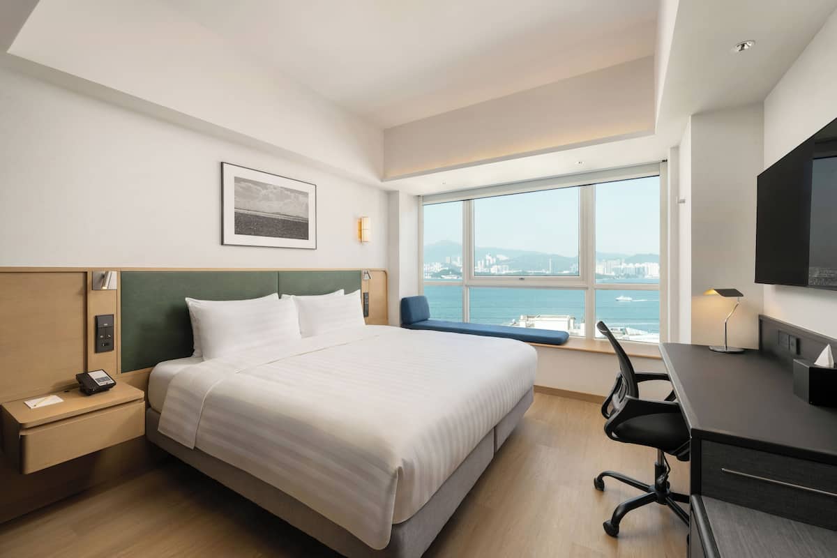 A hotel room with a bed, desk, chair, and a large window offering a city view.