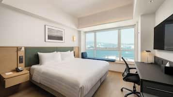 A hotel room with a bed, desk, chair, and a large window offering a city view.