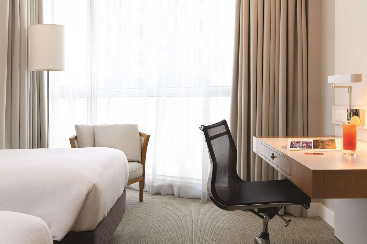 A hotel room with a bed, a desk with a chair, a lamp, and a window with curtains.